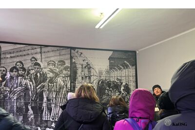 Grupa osób z tyłu w ciepłych ubraniach słucha prelekcji w jasnym pomieszczeniu. Za nimi znajduje się duża, czarno-biała grafika lub fotografia przedstawiająca grupę dzieci i młodzieży stojącej za drutami kolczastymi, prawdopodobnie więźniów obozu.