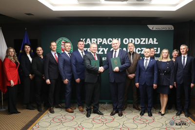 Na zdjęciu widać grupę ludzi stojących obok siebie. Na środku widać dwóch mężczyzn którzy trzymają w ręku zielone teczki i podają sobie ręce.