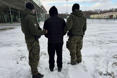 Zdjęcie od tyłu przedstawiające dwóch funkcjonariuszy Straży Granicznej eskortujących mężczyznę w czarnym ubraniu z kapturem na głowie. Funkcjonariusze są ubrani w zielone kurtki taktyczne i spodnie typu moro. Cała trójka stoi na zaśnieżonym terenie przed budynkami infrastruktury granicznej. W oddali widać zalesiony pas terenu pod zachmurzonym niebem.