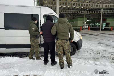 Dwaj funkcjonariusze Straży Granicznej w zimowych mundurach (oliwkowe kurtki i spodnie w kamuflażu) stoją przy białym samochodzie dostawczym na zaśnieżonym placu. Pomiędzy nimi znajduje się mężczyzna w czarnej kurtce z kapturem i czarnych spodniach, którego funkcjonariusze trzymają pod ramię. W tle widoczna jest wiata przejścia granicznego.