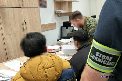 Zatrzymani dwaj obywatele Bangladeszu siedzą w pomieszczeniach służbowych placówki sg w tarnowie. jeden z funkcjonariuszy umundurowany  pochyla się nad dokumentami cudzoziemców leżącymi na biurku, drugiego funkcjonariusza w cywilnym stroju widać tylko ramię z odblaskową opaską z napisem straż graniczna