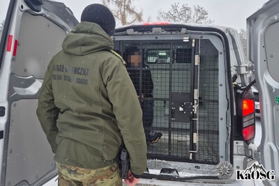 zatrzymany cudzoziemiec siedzi w samochodzie sg typu więźniarka. przed nim stoi tyłem funkcjonariusz SG uzbrojony i umundurowany