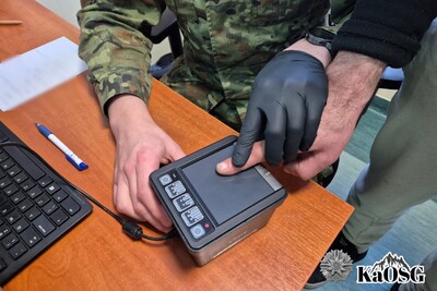 zbliżenie na biurko, na którym leży urządzenie do pobierania odcisków palców elektronicznie. Widać ręce funkcjonariusza w czarnych rękawiczkach, który pobiera odciski palców od zatrzymanego cudzoziemca