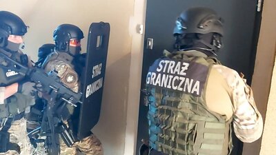 Uzbrojeni funkcjonariusze jednostki specjalnej Straży Granicznej w kaskach i kamizelkach taktycznych, z tarczą balistyczną, przygotowują się do wejścia do pomieszczenia.