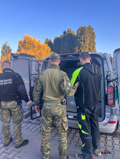 Na zdjęciu dwóch funkcjonariuszy SG przy drzwiach otwartej furgonetki służbowej. Jeden z nich trzyma pod ramię mężczyznę w uniformie pracowniczym. Najprawdopodobniej jest to osoba zatrzymana, którą wprowadzają do więźniarki.