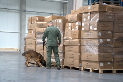Na zdjęciu mężczyzna w umundurowaniu specjalistycznym trzyma na smyczy psa. Pies sprawdza ustawione przed nim tekturowe pakunki w dużej ilości.