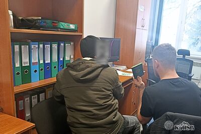 Na zdjęciu w środkowej jego części mężczyzna ubrany w kurtkę zimową koloru khaki oraz czarną czapkę zimową. Mężczyzna, prawdopodobnie zatrzymany, siedzi w pomieszczeniu służbowym a obok inny mężczyzna trzyma w ręku paszport.  Po lewej stronie na ścianie meble biurowe typu półki a na nich segregatory.