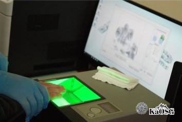 Zbliżenie na urządzenie do pobierania odcisków palców. Na czytniku widać dłoń zatrzymanej podtrzymywaną przez dłoń funkcjonariusza w rękawiczkach jednorazowych.