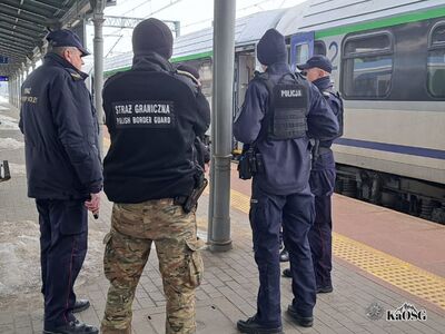 Na zdjęciu grupa funkcjonariuszy w umundurowaniu. Na pierwszym planie mężczyzna ubrany w spodnie moro i czarną bluzę z napisem STRAŻ GRNIACZNA, przy boku przypięta broń w kaburze, na głowie czapka. Z lewej strony mężczyzna w granatowym mundurze, na głowie czapka typu kaszkiet. z prawej strony inni mężczyźni w granatowych mundurach, jeden z nich ma przy boku broń i napis POLICJA. W tle fragment pociągu oraz dworca kolejowego.