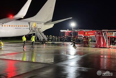 Na zdjęciu fragment samolotu pasażerskiego, na płycie lotniska. Zdjęcie zrobione w nocy. Widoczna płyta lotniska oraz osoby w zielonych kamizelkach, w tle samochody straży pożarnej oraz strażacy w roboczym umundurowaniu.