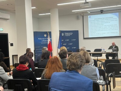 Zdjęcie w pomieszczeniu konferencyjnym. Przy biurku widoczny od przodu mężczyzna najprawdopodobniej prelegent.