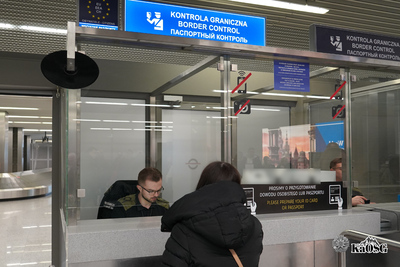 Przy stanowisku do kontroli na lotnisku stoi kobieta w czarnej kurtce. Za szybą w budce kontrolerskiej siedzi funkcjonariusz i dokonuje kontroli