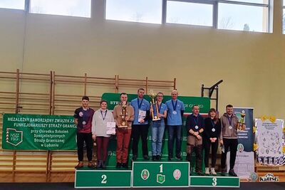 Na zdjęciu grupa osób , stojących prawdopodobnie w sali gimnastycznej na podium medalowym. Na każdym podium są trzy osoby, z których jedna trzyma w rękach puchar. W tle widoczne drabinki gimnastyczne oraz banery różnych organizacji i podmiotów.