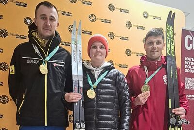 Na zdjęciu trzy osoby w sportowych strojach. Każda z nich ma zawieszony na szyi medal, dwie w rękach trzymają narty biegowe. W tle widoczny duży baner z logotypem i napisem Bieg Piastów.