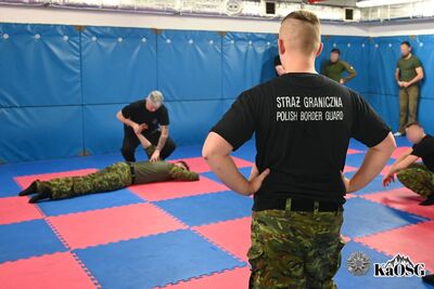 Widok zza pleców funkcjonariusza w koszulce „Straż Graniczna Polish Border Guard” na salę treningową. W tle instruktor pokazuje technikę interwencji osobie leżącej na czerwono-niebieskiej macie, a pozostali uczestnicy obserwują ćwiczenie.