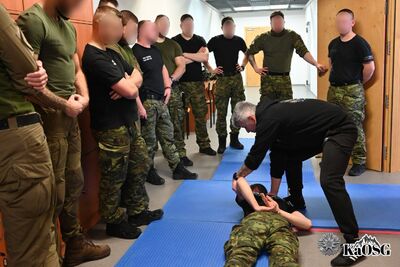 Szkolenie w węższym pomieszczeniu. Instruktor pochyla się nad leżącym na macie funkcjonariuszem, prezentując techniki zakładania kajdanek lub blokowania kończyn. Wokół stoi grupa kursantów w czarnych i oliwkowych koszulkach.