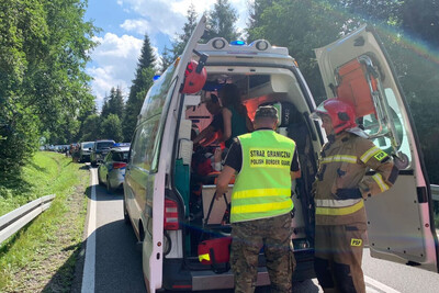 Poszkodowany leży w ambulansie. Lekarka udziela mu pomocy. Przed tylnymi drzwiami stoi funkcjonariusz Straży Granicznej, trzymający plecak ratowniczy oraz strażak