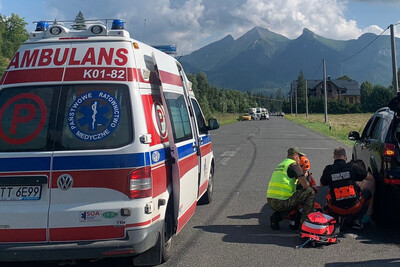 Na tle Tatr stoi ambulans. Funkcjonariusz Straży Granicznej i ratownik medyczny udzielają pomocy siedzącemu mężczyźnie z zawałem serca, opartemu o samochód osobowy.