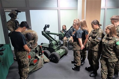 Uczniowie w mundurach w sali stoją i słuchają pogadanki. Funkcjonariusz prezentuje wizytującym robota pirotechnicznego.