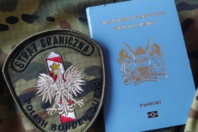 Służbowo z Kenii przez Hiszpanie do Polski emblemat straży granicznej z napisem straż graniczna polish border guard na środku emblematu znajduję się orzeł biały a za nim słupek graniczny białoczerwony. obok emblematu znajduje się paszport Republiki Kenii w kolorze niebieskim. emblematy oraz paszport znajdują się na materiale o wzorze  moro