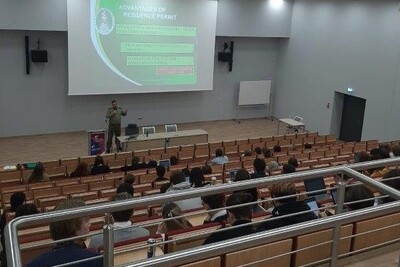 Sala wykładowa. Funkcjonariusz Straży Granicznej stoi przed siedzącymi w auli studentami - cudzoziemcami. W tle prezentacja w języku angielskim dot. zagadnień związanych z legalizacją pobytu na terytorium RP.
