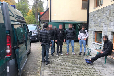 Zatrzymanych sześciu cudzoziemców stoi, a siódmy siedzi na ławce przed Placówką SG w Zakopanem.  Cudzoziemcy mają zapikselizowane twarze. Po lewej stronie i w tle samochody służbowe Straży Granicznej typu bus.