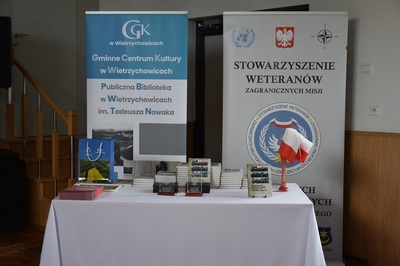 Na stole leży kilka stosów książek pt. Wspomnienia weteranów misji pokojowych i stabilizacyjnych z powiatu tarnowskiego. Przed nimi stoją dwie szklane statuetki z wizerunkiem orła. W tle dwa banery z napisem Gminne Centrum Kultury w Wietrzychowicach Publiczna Biblioteka w Wietrzychowicach im. Tadeusza Nowaka oraz  Stowarzyszenia Weteranów Zagranicznych Misji.