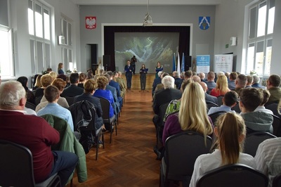 Sala, w której odbywa się premiera książki. Uczestnicy spotkania siedzą tyłem. W tle widać scenę i prowadzących uroczystość autorów publikacji.