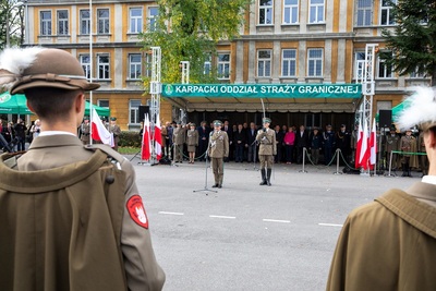 Komendant Karpackiego Oddziału Straży Granicznej witający się z zebranymi na uroczystości pododdziałami. Obok niego stoi dowódca uroczystości trzymający w ręku szablę. Za nimi trybuna honorowa z napisem Karpacki Oddział Straży Granicznej, na której znajdują się zaproszeni goście. Widoczne również flagi białoczerwone.