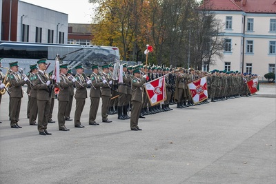 Na pierwszym planie tamburmajor Orkiestry Reprezentacyjnej Straży Granicznej który dyryguje orkiestrą za pomocą buławy trzymanej w ręku. W dalszej kolejności pododdziały zebrane na uroczystości. W tle pojazdy służbowe Straży Granicznej.