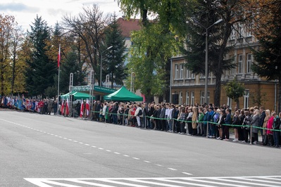 Na pierwszym planie widoczni uczestnicy uroczystości, nieco dalej trybuna honorowa, gdzie stoją zaproszeni goście. W dalszej perspektywie  poczty sztandarowe.