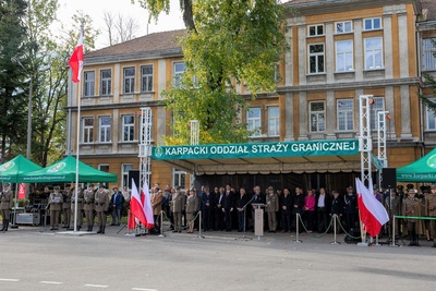 Trybuna honorowa z zaproszonymi na uroczystość gośćmi. Z lewej strony poczet flagowy składający się z trzech funkcjonariuszy Straży Granicznej. Na maszcie powiewa biało czerwona flaga Polski. Przed trybuną również znajdują  się powiewające biało-czerwone flagi.