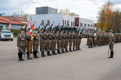 Kompania honorowa Straży Granicznej wykonująca salwę honorową. Przed kompanią stoi jej dowódca wydający polecenia. W tle pojazdy służbowe straży granicznej.