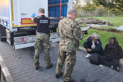 na pierwszym planie funkcjonariusz straży granicznej rozmawiający z zatrzymanymi cudzoziemcami. mężczyźni siedzą na krawężniku. drugi funkcjonariusz straży granicznej dokonuje oględzin naczepy tira, w którym podróżowali nielegalni migranci.