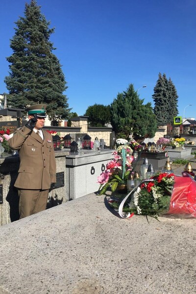 Cmentarz w Lubaczowie. Pułkownik Daniel Wojtaszkiewicz w umundurowaniu galowym stoi przed grobowcem patrona Placówki SG w Kielcach i oddaje honor zmarłemu. Na grobie leżą kwiaty, w  tym bukiet w biało-czerwonych barwach złożony przez Komendanta Placówki SG w Kielcach. W tle inne grobowce na miejscowym cmentarzu.
