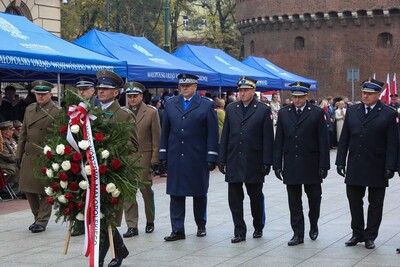 Siedmiu przedstawicieli służb mundurowych województwa idzie w szeregu by złożyć wieniec kwiatów. Przed nimi funkcjonariusz Straży Granicznej w historycznym mundurze niesie duży wieniec z biało-czerwonymi kwiatami. W tle Barbakan krakowski. Po lewej namioty z napisem Małopolski Urząd Wojewódzki w Krakowie, pod którymi stoją i siedzą zaproszeni na uroczystości rocznicowe goście.