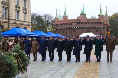Jedenastu przedstawicieli służb mundurowych województwa oddaje honor po złożeniu wieńca kwiatów. W tle Barbakan krakowski i uczestnicy uroczystości. Po lewej namioty z napisem Małopolski Urząd Wojewódzki w Krakowie, pod którymi stoją zaproszeni na uroczystości rocznicowe goście.