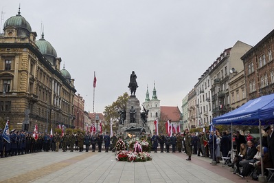 Plac Matejki w Krakowie. Pod pomnikiem stoi warta honorowa złożona z funkcjonariuszy służb mundurowych Małopolski. Przed pomnikiem leżą wieńce i wiązanki biało-czerwonych kwiatów. Na maszcie wywieszona jest biało-czerwona flaga, poniżej powiewają biało-czerwone flagi. Po lewej stoją poczty sztandarowe, po prawej namioty z napisem Małopolski Urząd Wojewódzki w Krakowie, pod którymi stoją i siedzą zaproszeni na uroczystości rocznicowe goście. W tle kamienice krakowskie oraz dwie wieże kościelne.