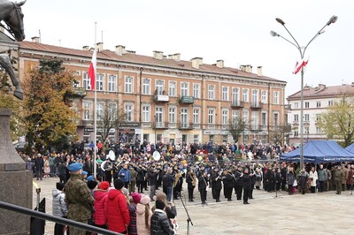 Plac Zamkowy w Kielcach. Na schodach przed kamienicami stoją zgromadzeniu uczestnicy uroczystości rocznicowych. Na placu gra orkiestra w mundurach, która gra hymn Polski. Ludzie stoją na baczność. Po lewej pod pomnikiem stoi schola dziecięca, za nią żołnierze pełnią wartę honorową pod pomnikiem.