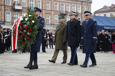 Kielce Plac Zamkowy. Trzech przedstawicieli służb mundurowych województwa świętokrzyskiego idzie w szeregu. Przed nimi funkcjonariusz Policji niesie duży wieniec z biało-czerwonymi kwiatami. W tle kamienice kieleckie i uczestnicy uroczystości.