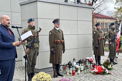 Nowy Sącz. Przed pomnikiem marszałka Józefa Piłsudskiego stoją funkcjonariusze Straży Granicznej oraz młodzież ze Strzelca pełniący wartę honorową. Pod pomnikiem złożone wiązanki biało-czerwonych kwiatów oraz znicze. Po lewej do mikrofonu przemawia moderator uroczystości.