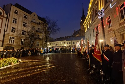 Tarnów. Ciemne niebo. Plac przed Grobem Nieznanego Żołnierza otaczają tarnowskie kamienice. W tle wieża katedry tarnowskiej. Po prawej stoją poczty sztandarowe. W oddali z przodu oraz po lewej stronie placu stoi duża grupa uczestników uroczystości.