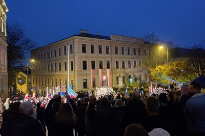 Tarnów. Ciemne niebo. Plac przed Grobem Nieznanego Żołnierza. W tle tarnowska kamienica. Na pierwszym planie stoi bardzo duża grupa uczestników uroczystości. Widać część pomnika Grób Nieznanego Żołnierza, powiewają biało-czerwone flagi.