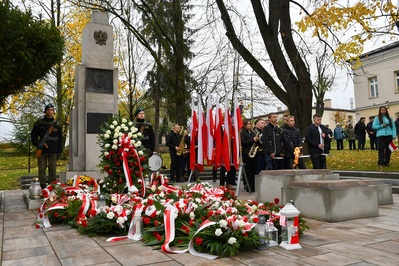 Dąbrowa Tarnowska. Pomnik Poległych Synów Miasta Dąbrowy, pod nim leży dużo wiązanek i stoi wieniec biało-czerwonych kwiatów. Powiewają flagi biało-czerwone. Młodzież w mundurach wystawiła wartę honorową. Po lewej stronie stoi młodzieżowa orkiestra. W tle uczestnicy uroczystości.