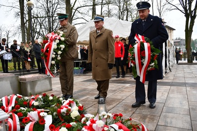 Dąbrowa Tarnowska. Przed pomnikiem Poległych Synów Miasta Dąbrowy, pod którym leżą wiązanki biało-czerwonych kwiatów, przedstawiciele służb mundurowych składają wieniec. Wśród nich Komendant Placówki SG w Tarnowie – kapitan SG Paweł Wolak.  W tle uczestnicy uroczystości.