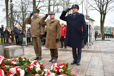 Dąbrowa Tarnowska. Przed pomnikiem Poległych Synów Miasta Dąbrowy, pod którym leżą wiązanki biało-czerwonych kwiatów, przedstawiciele służb mundurowych oddają honor. Wśród nich Komendant Placówki SG w Tarnowie – kapitan SG Paweł Wolak.  W tle uczestnicy uroczystości.