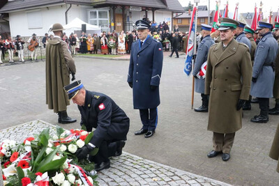 Rynek w Białym Dunajcu. Przed pomnikiem, pod którym leżą wiązanki biało-czerwonych kwiatów, przedstawiciele służb mundurowych składają wiązankę kwiatów. Wśród nich Komendant Placówki SG w Zakopanem – podpułkownik SG Andrzej Karkoszka.  W tle poczty sztandarowe oraz uczestnicy uroczystości, część w góralskich tradycyjnych strojach.