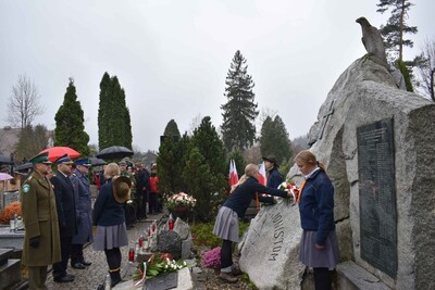 Cmentarz w Zakopanem. Przy Kwaterze Legionowej przedstawiciele służb mundurowych składają wiązankę biało-czerwonych kwiatów. Wartę przy grobie pełnia harcerki. W tle pod parasolami uczestnicy uroczystości.