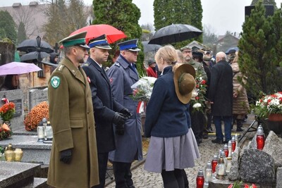 Cmentarz w Zakopanem. Przy Kwaterze Legionowej przedstawiciele służb mundurowych składają wiązankę biało-czerwonych kwiatów, które odbiera harcerka. W tle pod parasolami uczestnicy uroczystości.
