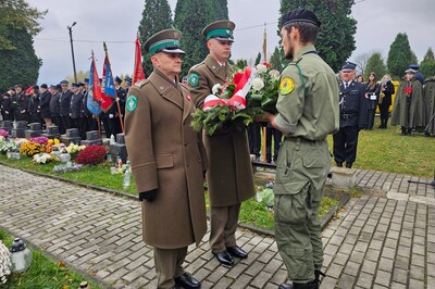 Cmentarz komunalny w Starym Sączu. Delegacja Karpackiego Oddziału Straży Granicznej przekazuje harcerzowi wieniec biało-czerwonych kwiatów. W tle poczty sztandarowe i uczestnicy uroczystości.